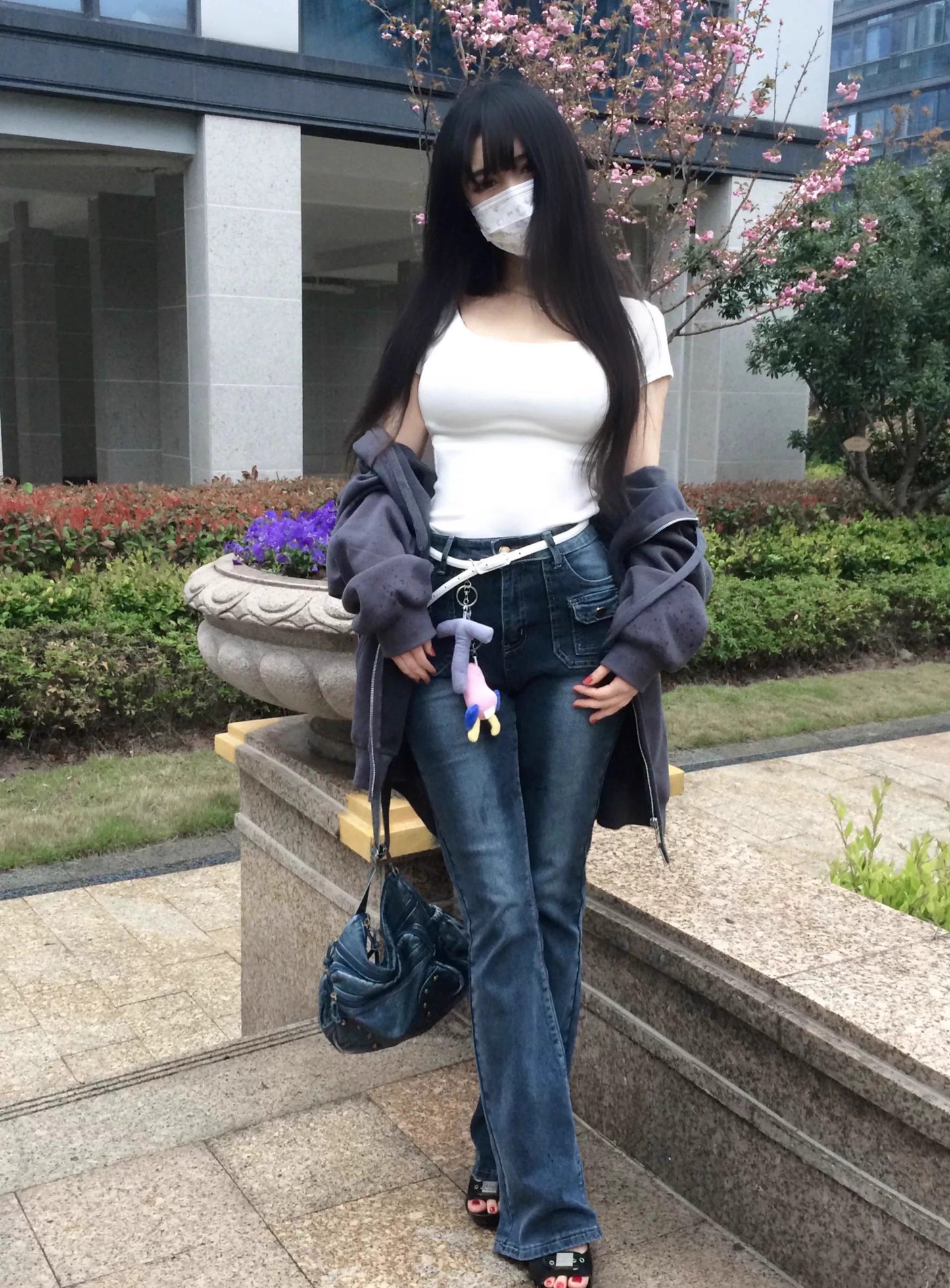 Person wearing a white top and blue bag standing outdoors with a building and tree in the background.