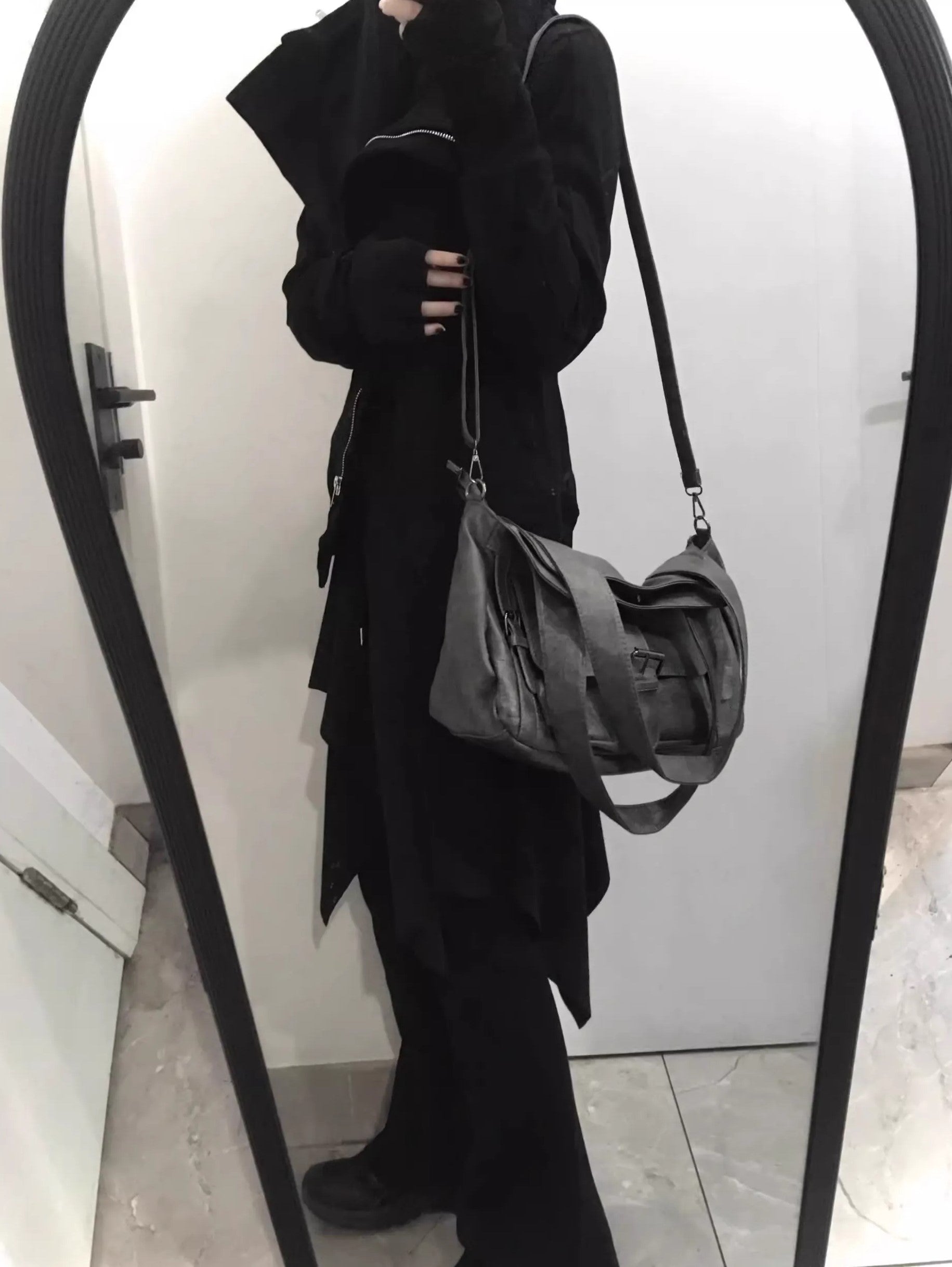 Person wearing a black outfit with a gray bag in front of a mirror.