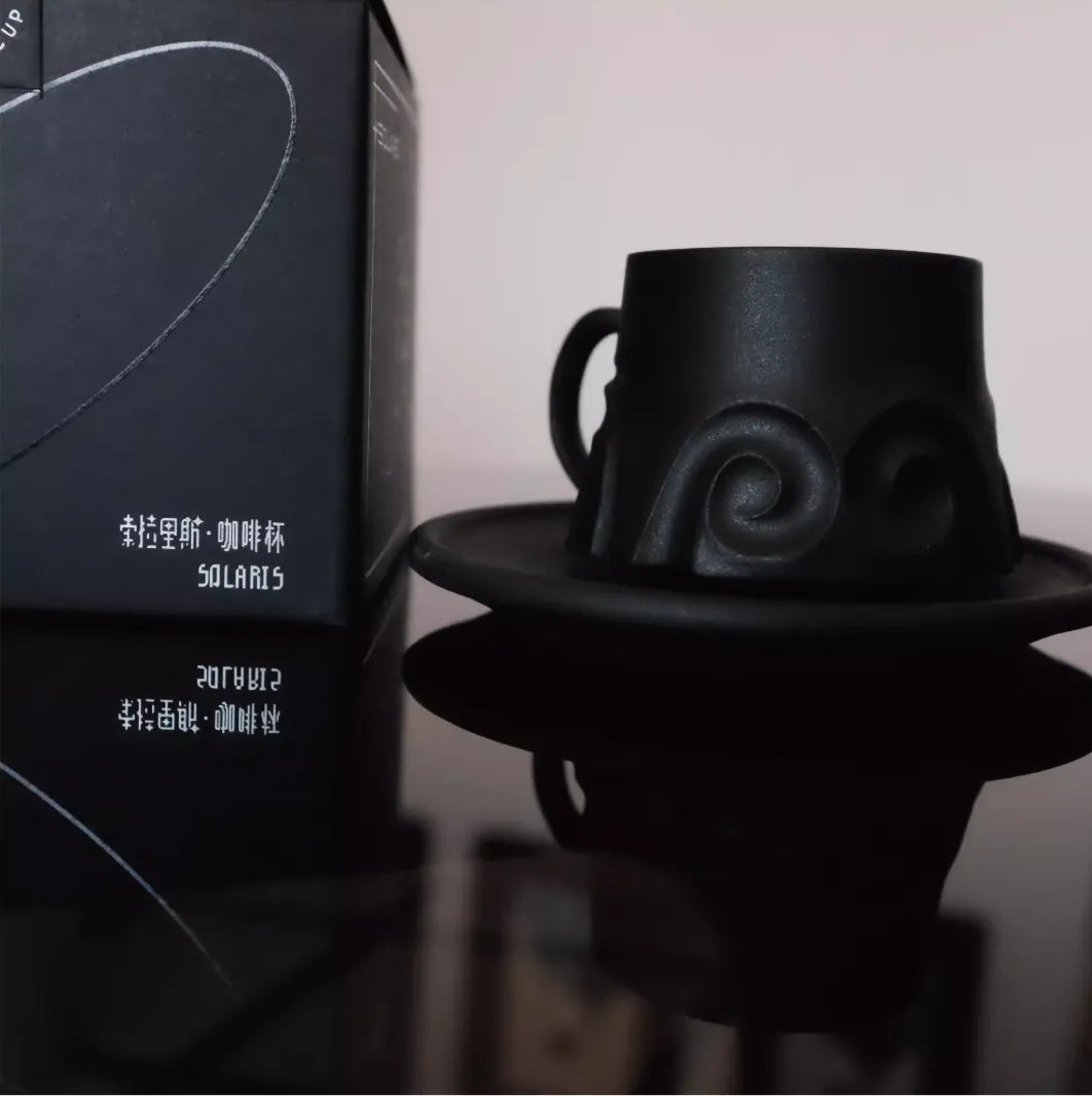 Black ceramic cup and saucer with a unique design, placed on a reflective surface next to a branded box.