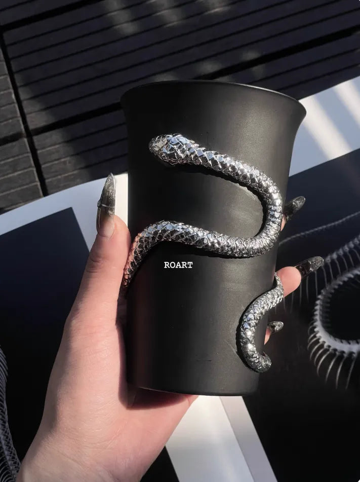 Black cup with a silver snake design held by a hand, on a wooden surface.
