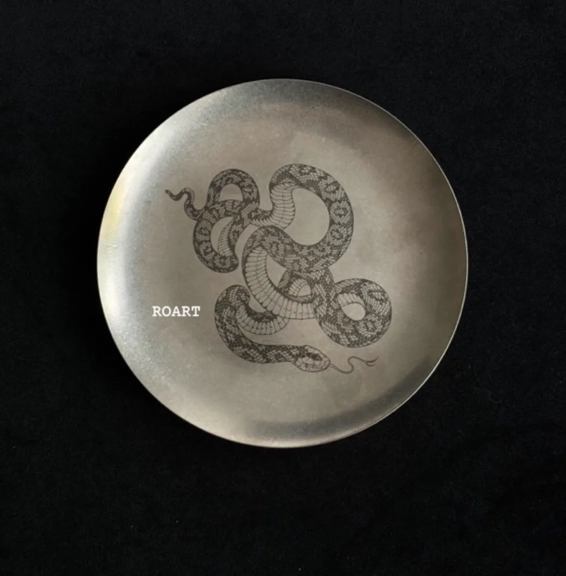 Round metallic plate with snake design and 'ROART' branding on a black background