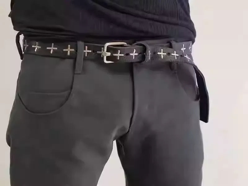 Person wearing black pants with a belt featuring white cross patterns on a plain background